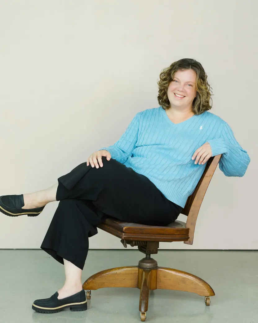 Alix Horton sitting sideways in a wooden swivel chair with her legs crossed, smiling, in a light blue sweater and black trousers against a plain pale wall.