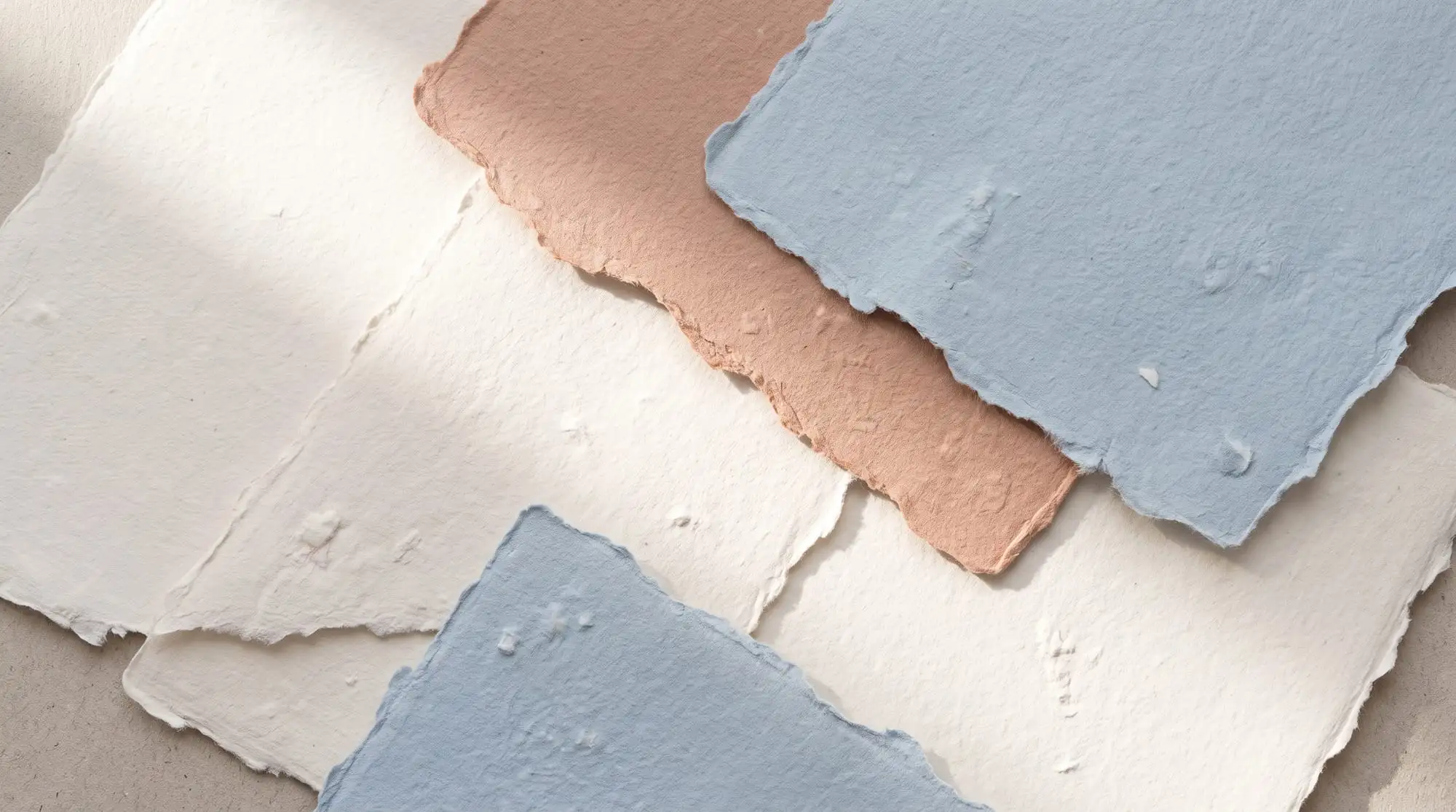 Overlapping sheets of torn handmade paper in cream, blush and soft blue, lit at a diagonal.
